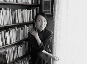 Photo of Author Kyo Maclear beside a bookshelf full of books and a bright window